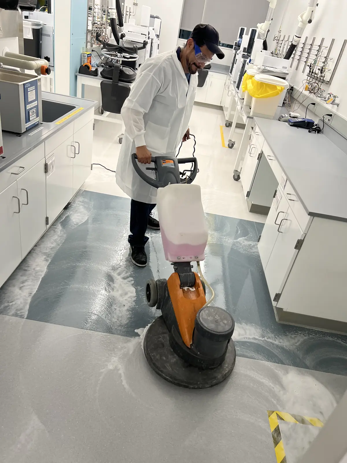 Technician in lab coat and PPE running a floor machine between lab benches