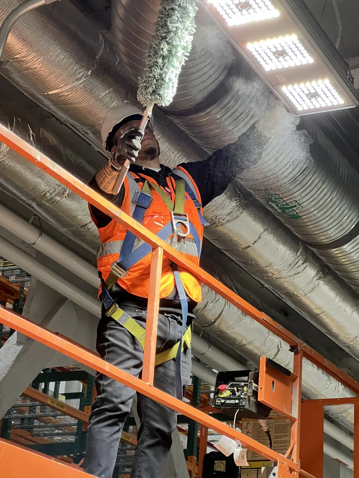 Nexara crew member in a safety harness dusting overhead ductwork from a lift